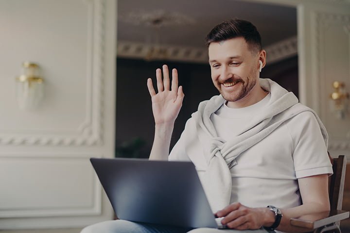 portrait of businessman or freelancer greeting somebody via laptop while sitting on chair at home